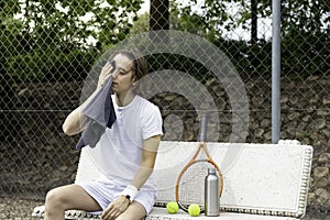 Young man wiping his sweat with a towel sitting on a bench after playing a game of tennis on a court