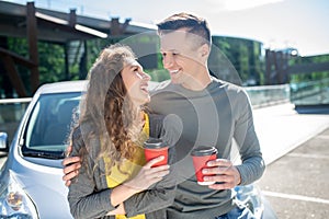 Young man and woman with coffee near a car.