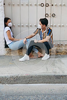 A young man and woman in a disposable mask dressed casually talking while sitting outdoors