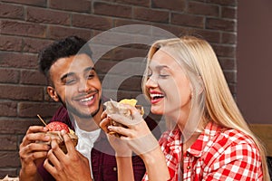 Young Man And Woman Eating Fast Food Burgers Sitting At Wooden Table In Cafe