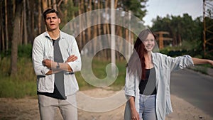 Young man and woman gesturing hitchhiking on suburban road at forest standing outdoors. Portrait of handsome boyfriend