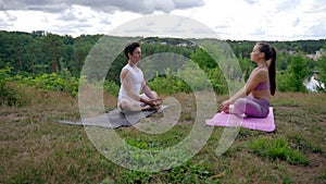 Young man and woman meditate in Lotus poses on hilly bank