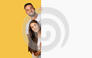 A young man and woman peek around a blank white sign with joyful expressions