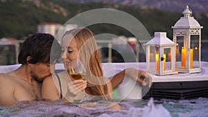 A young man and woman are relaxing in the hot tub on a rooftop with a view on mountains during sunset