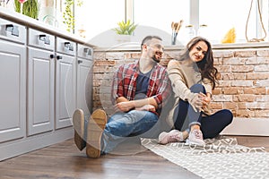Young man and woman sitting on floor in kitchen and talking. Loving young couple spending time together at home