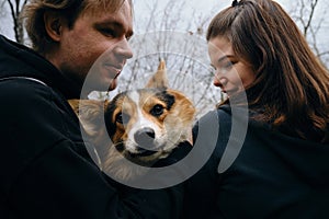 A young man and woman are smiling and hugging with their cute Welsh corgi puppy in the park. Dog with happy expression