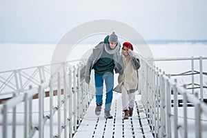 A young man and woman spend time together on a winter vacation, walking on a snow-covered pier on the river