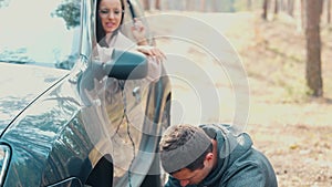 Young man and woman stand together in forest. Guy inflate car tires with air using compressor. Girl sit inside car and