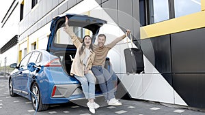 Young man and woman traveling by electric car having stop at charging station. Boyfriend and girlfriend sitting in the car trunk