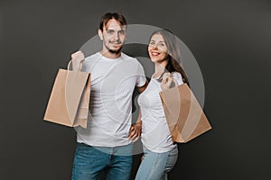 Young man and woman in white blank T-shirts with paper bags in their hands on a solid gray background. Studio photo