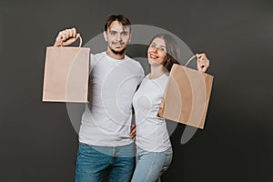 Young man and woman in white blank T-shirts with paper bags in their hands on a solid gray background. Studio photo