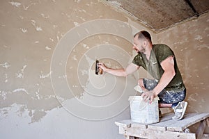 Young man at wooden stand is puttying the walls indoors. Guy with beard in t-shirt and jeans is smeared with putty photo