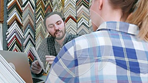 Young man worker chatting with customer about picture frame details, using laptop computer on counter in atelier