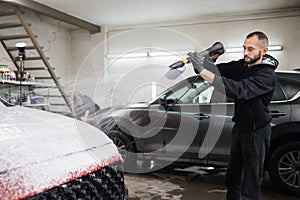 Young man worker cleaning modern red car hood with foam and pressurized water at service station