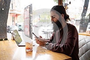 young man working from cafe using smart phone and laptop computer, hipster guy using smartphone in interior coffee shop