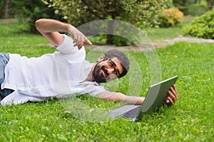 Young man working on his laptop while lying on the grass of the park.