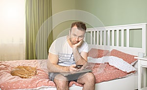 Young man working from home in bedroom, feelling bored and tired holding a phone and laptop in his arms
