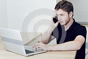 Young man working at home on a laptop talking on a cell phone, sitting at desk.