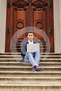 Young man working on laptop computer, sitting on stairs  outside office building in New York City