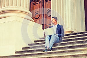 Young man working on laptop computer, sitting on stairs  outside office building in New York City