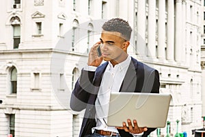 Young man working on laptop computer, talking on cell phone outside office building in New York City