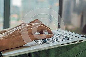 Young man is working on a laptop in his bed on a background of a panoramic window overlooking the skyscrapers