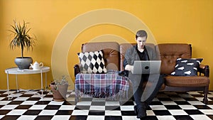 Young man working on laptop at home. Stock footage. Guy is working at home as freelancer on laptop sitting on couch