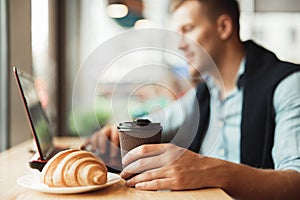 Young man working remotely in his laptop eats croissant and holds hot coffee in cafe