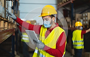 Young man working in warehouse doing inventory using digital tablet and loading delivery boxes plan while wearing face mask