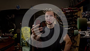 Young Man In Workshop Talking On The Phone. He Shows Himself On Camera. Cheerful Man Talking And Smiling.