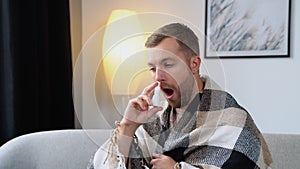 A young man wrapped in a blanket sprays a nasal spray while sitting on sofa
