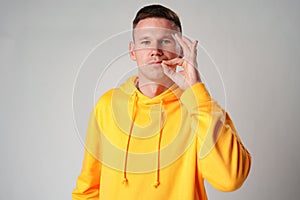 Young man in yellow hoodie making silence gesture against gray background