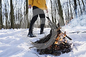 Young man in yellow jacket in the winter forest standing near small fire.