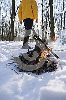 Young man in yellow jacket in the winter forest standing near small fire.