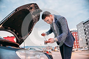 Young manager formally dressed checking his car oil at sunset while leaning under the car`s hood