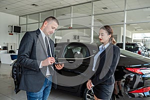 young manager in suit holding clipboard with female client talking to sales modern car