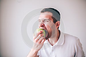 Young manin white shirt isolated over background. Guy biting piece of green fresh tasty apple. Delicious yummy fruit.