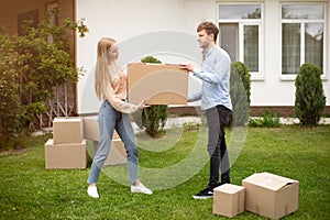 Young married couple carrying cardboard box in front of their new home on moving day