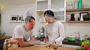 Young married couple cooking together in kitchen at home. Playful woman teasing and feeding her man with kiwi and fruits