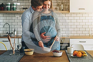 Young married couple embraces standing near table in kitchen. Husband hugs his pregnant wife