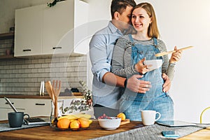 Young married couple embraces standing near table in kitchen. Husband hugs his pregnant wife