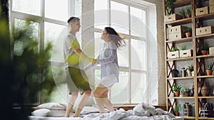 Young married couple is jumping and dancing on double bed in light room with large windows, happy people are having fun