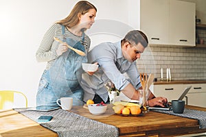 Young married couple in kitchen. Man stands near table and uses laptop, pregnant wife is standing next to him