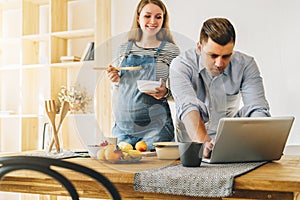 Young married couple in kitchen. Man stands near table and uses laptop, pregnant wife is standing next to him