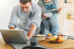 Young married couple in kitchen. Man stands near table and uses laptop, pregnant wife is standing next to him