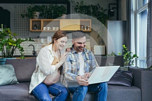 Young married couple man and pregnant woman at home on sofa using laptop for online shopping and video communication