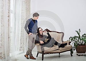 Young married couple posing on brown classy sofa in white room with big windows and tulle curtain. Romantic relationship. Happy