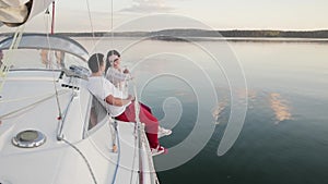 Young married couple in red pants sits on the side of a white sailing yacht sailing through dark calm water