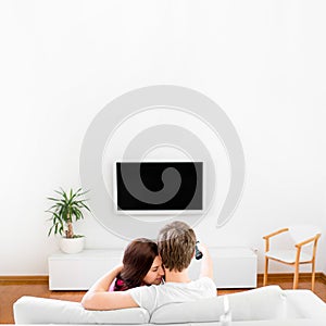 Young married couple sitting on the couch and watching tv at home, rare view, with space for text