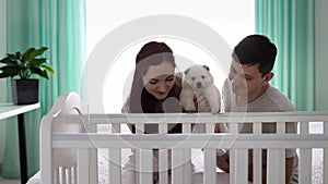 A young married couple with a small white puppy are looking into the crib where the newborn is sleeping. The concept of